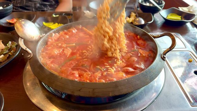 Footage of a hand cooking budae jjigae or Army base stew, South Korean food, on a hot pot. Seen here the noodles being cooked, with the smoke vapor flying up from the stew, with some gochujang paste.