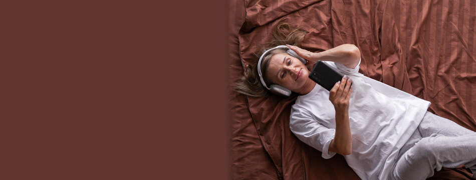 leisure technology. Happy middle aged woman with wireless headphones using smartphone in bed, while relaxing in bedroom with mobile phone, listening to music