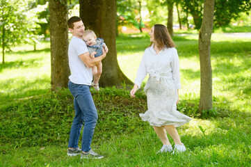 Fototapeta premium In public park, man and woman lovingly hold tiny baby in their arms, surrounded by bright green trees and grass, having fun, dancing, spinning. Summer fun for families in nature