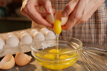 A person cracking an egg into a glass bowl, with several eggs in an egg carton in the background. The scene is set in a kitchen, showcasing the process of preparing food.