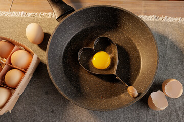 Heart-shaped frying pan for scrambled eggs with broken egg on kitchen table background, cardboard...