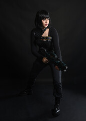 Full length portrait of beautiful black female model wearing sci fi futuristic dystopian leather bodysuit costume. Standing pose, holding gun weapon. isolated dark studio background moody silhouette.