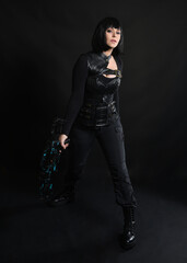 Full length portrait of beautiful black female model wearing sci fi futuristic dystopian leather bodysuit costume. Standing pose, holding gun weapon. isolated dark studio background moody silhouette.