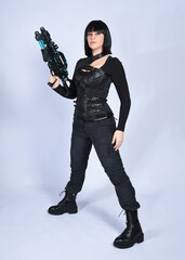 Full length portrait of beautiful black female model with short black hair wearing sci fi futuristic dystopian leather bodysuit costume. Standing pose holding gun weapon, isolated white studio backgro