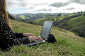 A scenic image of a person sitting on a grassy hillside, using a laptop computer with a view of rolling green hills in the background, evoking a sense of peace and productivity.