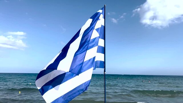Bandiera della Grecia che sventola sulla riva dell'isola di Rodi, con il mare sullo sfondo e la spiaggia con gli ombrelloni