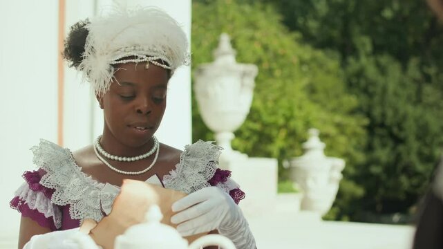 Zoom in shot of young African American aristocratic woman in feathered hat, gloves and Victorian dress receiving paper letter and reading it with smile during teatime outdoors