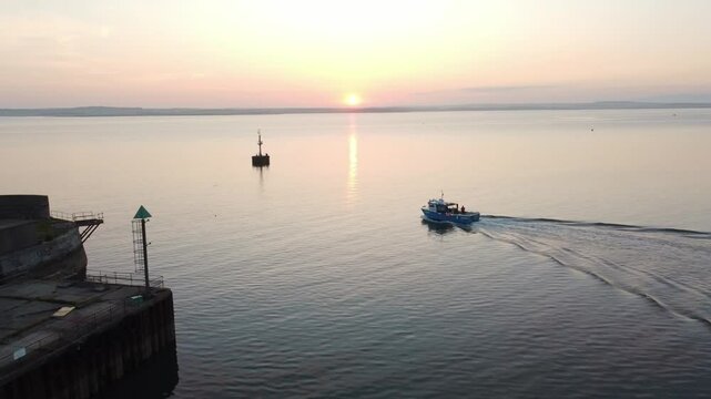 Small fishing boat passing old Holyhead lighthouse leaving wake ripples in sunlit sunrise seascape