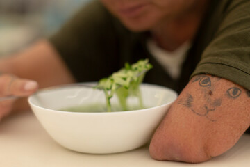 A person casually resting their arm on a wooden table next to a steaming bowl of fresh broccoli soup