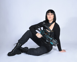 Naklejka premium Full length portrait of beautiful black female model, black hair wearing sci fi futuristic dystopian leather bodysuit costume. sitting pose, holding gun weapon, isolated white studio background.