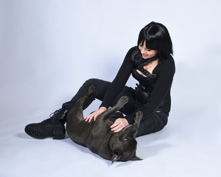 Full length portrait of beautiful black female model wearing sci fi futuristic dystopian leather bodysuit costume, with animal dog pet companion. sitting pose, isolated white studio background.