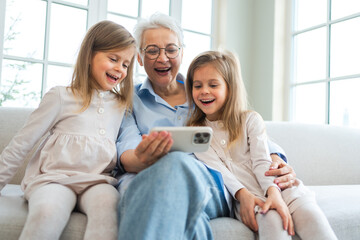 Happy family at home. Two little girls sisters twins grandmother enjoying time together watching video on phone. Good time at home. Grandma granddaughters child kids emotional bonding hugging together