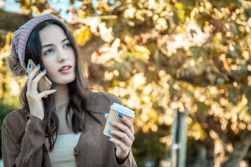 Woman talking on phone while drinking takeaway in park in autumn