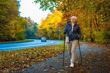 Beautiful middle-aged blonde woman in casual wear walking with poles on sidewalk by street in autumn scenery. Front view. Autumnal Nordic walking in city.