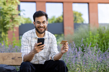 Fototapeta premium A smiling Indian man in glasses and a shirt is sitting on a bench outside, holding a phone and using a credit card