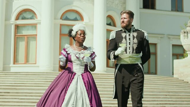 Young Black woman in period dress, feathered fascinator and gloves talking with Caucasian man in ornate uniform while walking outdoors in front of palace and having drinks