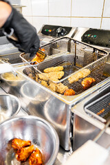 Close-up of a gloved hand placing breaded chicken pieces into a deep fryer basket in a commercial kitchen. The scene captures the preparation process in a professional culinary environment.