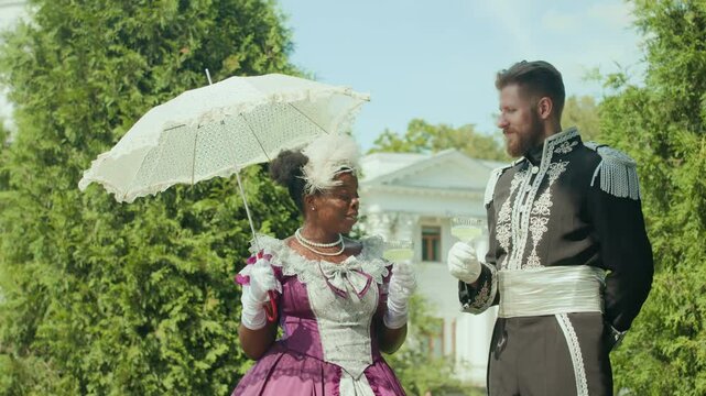 Young aristocratic Black woman in vintage dress holding lace parasol and drink in glass, smiling and chatting with Caucasian officer in ornate uniform outdoors on sunny day