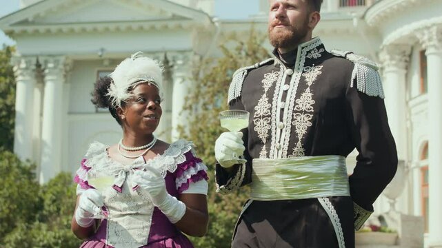Caucasian nobleman in ornate uniform holding glass of refreshing drink and chatting with African American aristocratic lady while strolling together outdoors on sunny summer day