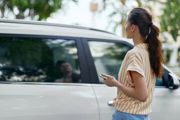 Woman standing near a van, holding a remote, contemplating, outdoor setting with greenery, casual attire, sunlight reflections, urban environment, summer vibes, vehicle parking scene © SHOTPRIME STUDIO