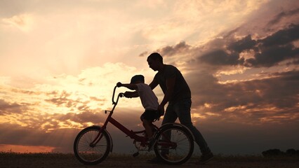 Fototapeta premium happy family in the park. father teaching son to ride a bike at sunset silhouette in the park. son child learning to ride a bike at sunset father helping son. child playing riding lifestyle a bike