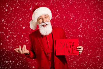 Photo of positive funny santa claus hold sale placard cheerful smile wear x-mas hat suit on red color background