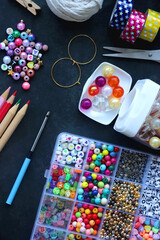 Colorful beads, coloring pencils, glitter, needles, string, hoop, tape and scissors on dark background. Various colorful craft supplies. Flat lay.