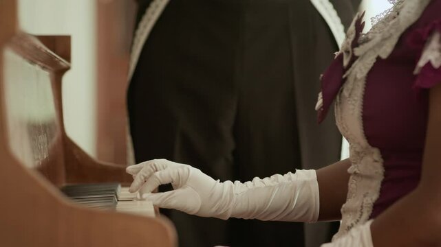 Close up view of hands of aristocratic woman wearing elegant white gloves and vintage dress playing grand piano while officer in uniform standing beside