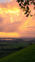 Peaceful sunset landscape featuring orange-pink clouds, sunrays illuminating a quiet countryside, distant town, serene lake, green fields, and soft light over a grassy hill with trees.