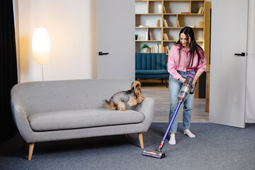 Cute young woman vacuuming at home with a cordless vacuum cleaner while her cute dog watches.