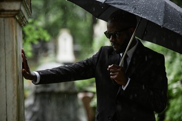 Medium shot of adult Black man in funeral suit mourns loss of loved one touching memorial monument...