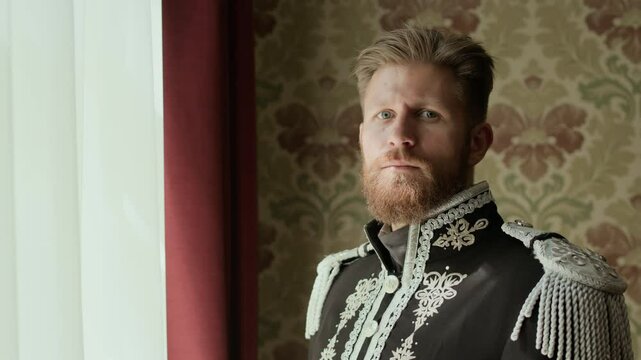 Zoom in portrait shot of bearded officer wearing ceremonial dress uniform with ornate embroidery standing in living room, looking through window and posing for camera with confidence