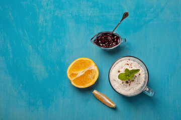 A light dessert featuring whipped cream topped with a mint garnish in a glass, accompanied by a halved orange, a slice of orange, and a small glass dish of blueberry jam on a vibrant blue background.