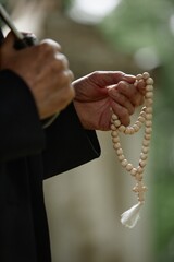 Vertical shot of hands of unrecognizable senior person holding wooden rosary beads praying for soul of deceased at cemetery while holding umbrella, copy space