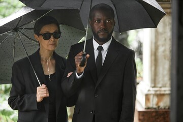 Medium shot of adult Black man in funeral suit standing by senior wife in sunglasses grieving loss...