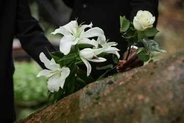 Close up on rain wet white lilies and rose in hands of two unrecognizable people placing flowers on...