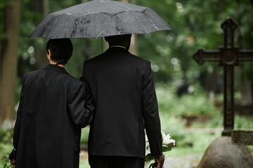 Rear view of senior woman with Black man by hand walking under umbrella along graves at cemetery...