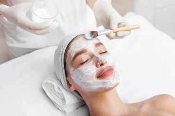 Beautician applying clay mask on face of young woman, lady enjoying beauty treatments in spa salon,...