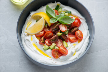 Bowl of greek feta dip topped with fresh tomatoes and cucumber, horizontal shot on a grey granite background, selective focus