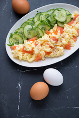 Scramble with smoked salmon and cucumber salad, vertical shot on a black marble background, elevated view