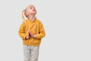 Blonde Caucasian little girl in yellow hoodie and gray pants standing with folded arms, looking up,...