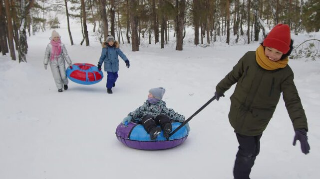 Tracking shot of smiling and happy diverse little boys and girls in warm winter clothes going up hill to ride on inflatable sledge snow tubes during winter vacation. Boy pulling girl up hill on sled