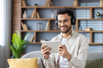 A young Indian man wearing headphones is sitting on the sofa at home, holding a tablet and looking at the screen with a smile