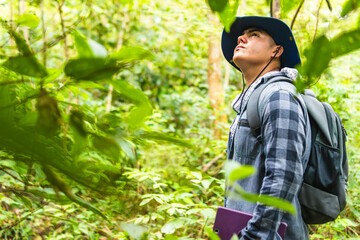 Young Latino biologist with a bag and a tablet, looking up at the upper parts of a forest where he is conducting research or work on the environment and Amazon rainforests