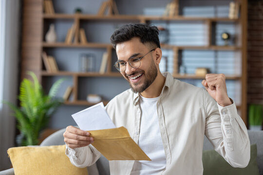 Close-up photo of a young indian man in a shirt and bib sitting on the couch at home and reading a letter he received, happy with the news and message