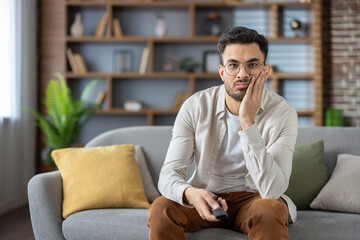 Tired and bored young indian man sitting in front of camera at home, holding head in hand and holding tv remote control