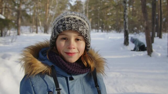 Chest up portrait of positive Caucasian boy in beanie and blue parka posing in winter forest on warm day and looking at camera, his friends running in background