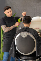 tattooed barber cutting the hair and beard of a young, dark-haired client.