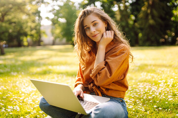 A young woman enjoys a sunny afternoon in the park, engaging with her laptop while sitting comfortably on the grass surrounded by nature and flowers. Freelancer, Online education.