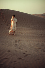 Woman in white garment walking on desert dune. Golden light enhances mood. Footprints in sand tell story of journey. Conveys solitude, adventure, and cultural richness.